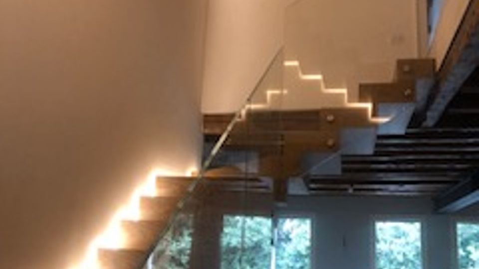 Interno moderno con scala sospesa in legno e vetro illuminata da strisce LED lungo i gradini, sotto un soffitto alto con travi a vista. Al piano inferiore si vede un’area soggiorno con poltrone e grandi finestre che affacciano sul verde esterno.