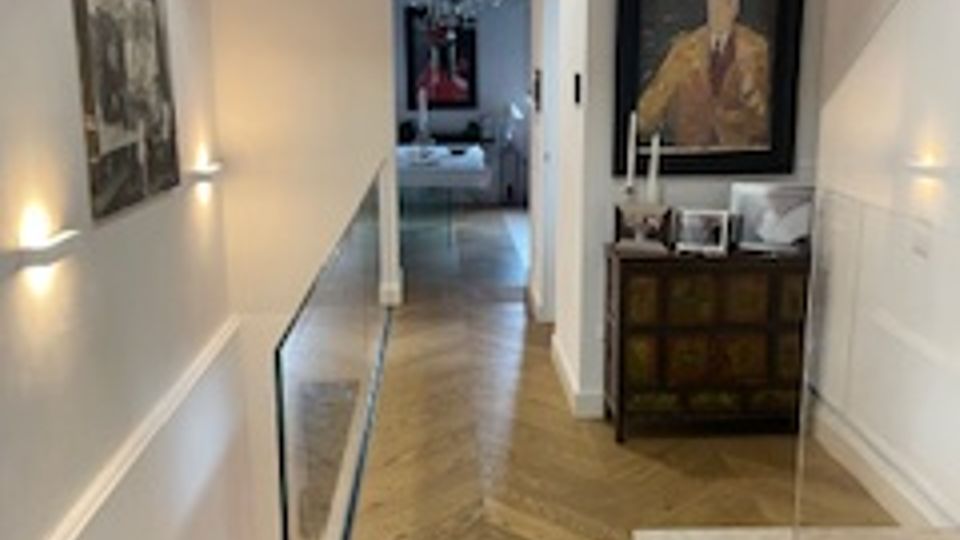 Corridoio interno di una casa con pareti bianche, soffitto con travi in legno a vista e pavimento in parquet a spina di pesce. Lungo le pareti ci sono applique, quadri e una credenza con un grande dipinto, con un’apertura verso un’altra stanza in fondo.