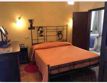 Camera da letto con letto matrimoniale in ferro battuto e copriletto arancione, illuminata da appliques a parete e con pavimento in parquet. Sulla destra una porta aperta conduce a un piccolo bagno con lavabo e asciugamani visibili.