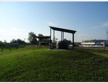 Prato erboso con piccole strutture moderne in metallo e copertura inclinata, che riparano volumi arrotondati scuri su basi in cemento. Sullo sfondo si vedono alberi, una strada con guardrail e edifici industriali sotto un cielo sereno.