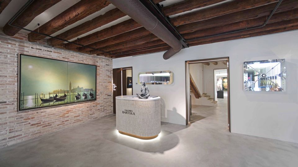 Interno di una galleria d’arte moderna con pavimento in cemento, pareti bianche e una parete in mattoni a vista con grande opera esposta. Al centro c’è un banco reception curvo illuminato, mentre il soffitto presenta travi in legno e tubazioni a vista con ingressi verso altre sale.