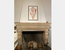 Grande camino in pietra con ampia cappa e focolare annerito, con alcuni utensili e recipienti in metallo e terracotta davanti. Sopra la mensola è appeso un pannello con uno stemma araldico su parete bianca.