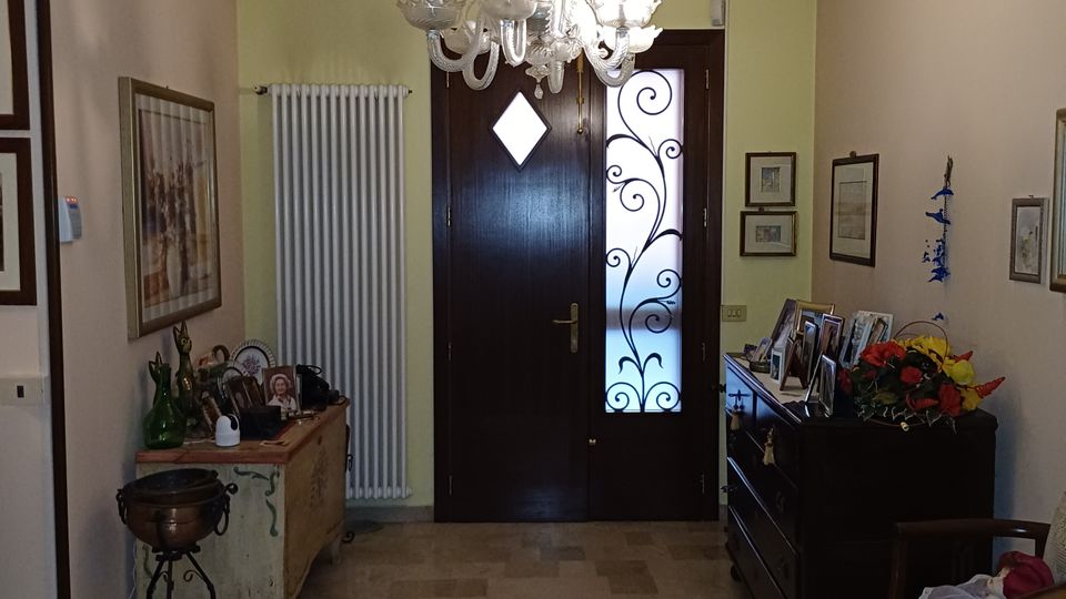 Ingresso di abitazione con pavimento in pietra chiara, lampadario in vetro al soffitto e porta d’ingresso in legno scuro con inserto in vetro decorato. Lungo le pareti ci sono un radiatore verticale, mobili bassi con cornici e oggetti decorativi e quadri appesi.