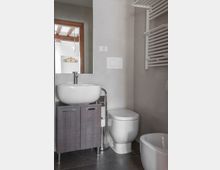 Bagno moderno con pareti piastrellate grigie, lavabo ovale su mobile, specchio con cornice in legno e WC con placca di scarico a parete. A destra si vedono un termoarredo bianco e il bordo di una vasca.