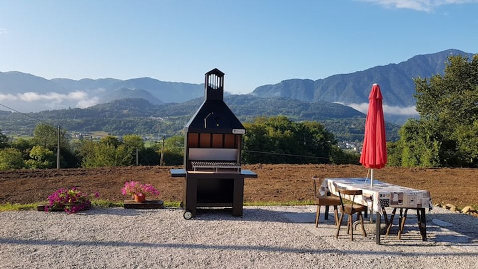 Terrazza in ghiaia con barbecue in muratura al centro e tavolo da esterno con sedie e ombrellone rosso sulla destra. Sullo sfondo si vede una vallata con alberi e campi e una catena montuosa sotto un cielo sereno.
