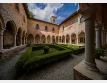 Chiostro storico in mattoni con porticato ad archi su colonne in pietra che circonda un giardino centrale con siepi geometriche e vialetti in ghiaia. Sulle pareti si vedono finestre ad arco e affreschi, con un piccolo campanile che sporge sopra i tetti.