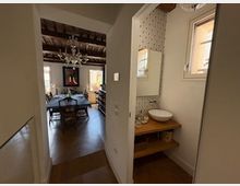 Interno di una casa con corridoio che conduce a una zona pranzo con tavolo e sedie, soffitto con travi a vista e lampadario. In primo piano c’è un piccolo bagno con lavabo su mensola in legno, parete a piastrelle decorate e una finestra.
