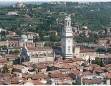 Veduta panoramica di una città collinare con tetti in terracotta, dominata da una grande chiesa in pietra a fasce e da un alto campanile quadrato. Sullo sfondo si vedono pendii verdi con cipressi, ville sparse e un edificio monumentale su una collina.