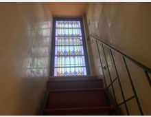 Vano scala interno con gradini in legno e corrimano in metallo, che conduce a una grande finestra verticale con vetrata colorata a motivi geometrici. Pareti lisce e alte, illuminate dalla luce naturale filtrata dalla vetrata.