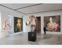 Interno di una galleria d’arte contemporanea con pareti bianche e pavimento grigio, illuminata da un soffitto a pannelli. Nella sala sono esposti grandi quadri figurativi alle pareti e una scultura su piedistallo al centro, con una vetrata laterale che affaccia su un edificio in mattoni.