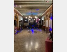 Salone interno in stile storico con soffitto a travi dipinte, lampadario centrale e pareti decorate con archi e specchi dorati. La sala ha pavimento in terrazzo con medaglione centrale ed è allestita per una festa con palloncini, luci colorate e postazione audio sul fondo.