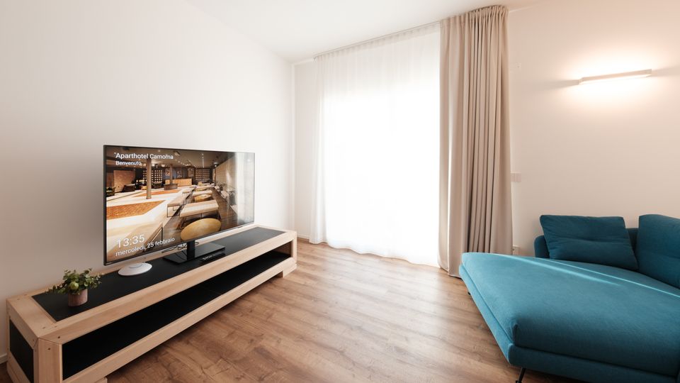 Soggiorno moderno e minimalista con pavimento in legno, pareti bianche, mobile TV basso con televisore e grande finestra con tende chiare fino a terra. A destra si vede un divano angolare blu sotto una lampada a parete.