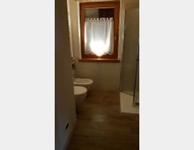 Bagno moderno con pavimento effetto legno, sanitari bianchi sospesi e box doccia in vetro sulla destra. Sul fondo c’è una finestra con cornice in legno e tendina bianca, sopra pareti rivestite con piastrelle chiare.