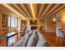 Ampio soggiorno di un appartamento con soffitto a travi in legno a vista, pavimento in parquet e illuminazione a incasso. Nella stanza si vedono una libreria a parete, finestra con inferriata decorativa, divano e poltrona, tavolino, quadri alle pareti e un’area pranzo con tavolo e sedie.