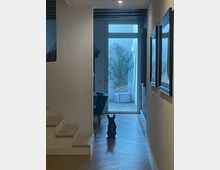 Corridoio interno con pavimento in legno che conduce a una porta a vetri, oltre la quale si vede un piccolo patio con una sedia e piante. A sinistra ci sono gradini e una parete divisoria, mentre a destra sono appesi specchi rettangolari lungo la parete.