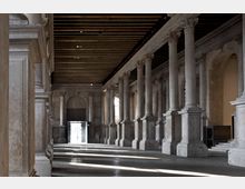 Ampio portico interno in pietra con lunga fila di colonne e archi, soffitto a travi in legno e pavimento in marmo. In fondo si vede un grande portale che lascia entrare luce naturale, creando fasci luminosi sul pavimento.