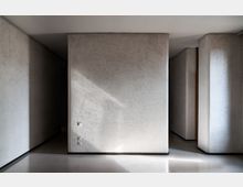 Interno di un corridoio moderno con pareti spesse intonacate in grigio chiaro, volumi rettangolari e angoli arrotondati. Pavimento lucido e chiaro con luce naturale che crea ombre morbide lungo i passaggi laterali.