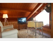 Interno di una mansarda in legno con soffitto spiovente e travi a vista, arredata con divano chiaro, lampada da terra e televisore su tavolino. Accanto a una grande finestra con tende c’è un tavolo da pranzo con tovaglia e sedie, su un tappeto decorato.