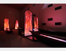 Interno di una mostra d'arte contemporanea con pareti in tonalità scura, caratterizzato da grandi installazioni verticali illuminate da luci rosse e arancioni simili a fuoco, e barche scure disposte sul pavimento.