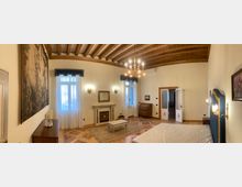 Ampia camera da letto in stile classico con soffitto a travi di legno a vista, pavimento in parquet a intarsi e lampadario centrale. La stanza include un letto con testata decorata, due finestre con tende leggere, un camino ornamentale, un arazzo a parete e una porta a doppio battente che conduce a un’altra stanza.