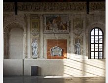 Interno di una sala storica con parete affrescata, lesene e archi decorativi, con riquadri pittorici e figure in stile classico ai lati di una lapide centrale. A destra una grande finestra ad arco con grata illumina il pavimento con fasci di luce.