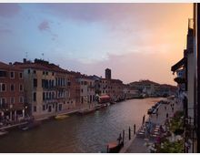 Veduta di un canale cittadino al tramonto, con edifici storici dai tetti in tegole e facciate con finestre ad arco lungo le rive. Sul canale si vedono piccole imbarcazioni e pontili, mentre a destra corre una passeggiata con tavolini all’aperto e balconi fioriti.