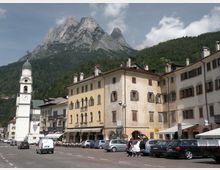 Piazza di un paese alpino con palazzi storici color pastello e portici al piano terra, affacciati su un’area di parcheggio. A sinistra si vede un campanile bianco con orologio, sullo sfondo una grande montagna rocciosa e un versante boscoso sotto un cielo parzialmente nuvoloso.