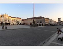 Ampia piazza cittadina lastricata in pietra, circondata da edifici storici con portici e facciate color pastello. Al centro si vede un alto pennone su basamento e, sullo sfondo, una torre civica con orologio sotto un cielo sereno.
