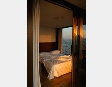 Camera da letto moderna con letto matrimoniale e pavimento in legno, illuminata da luce calda, con tende chiare e parete con testiera in legno. Una grande porta-finestra aperta affaccia su un tratto d’acqua, visibile all’esterno.