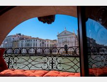 Vista da una terrazza coperta con ringhiera in ferro battuto sul Canal Grande, con facciate di palazzi storici e una chiesa monumentale in pietra sull’altra sponda. In primo piano si vedono cuscini rossi e una vetrata laterale che riflette parte della scena.