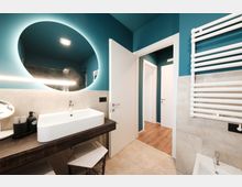 Bagno moderno con pareti blu petrolio e rivestimento in piastrelle grigie, lavabo rettangolare su mensola in legno scuro e grande specchio rotondo retroilluminato. A destra si vede un termoarredo bianco e, sul fondo, la porta aperta verso un corridoio con pavimento in legno.