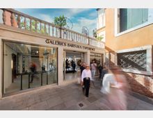 Ingresso di una galleria d’arte con ampia vetrata e portale centrale, insegna “Galeries Bartoux Experience” sopra la facciata chiara con balaustra in pietra. La scena è in una stretta calle lastricata tra edifici dai muri intonacati e in mattoni, con dettagli di finestre e grate.