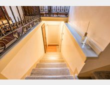 Vano scala interno illuminato, con gradini in pietra che scendono verso un pianerottolo in legno e porte laterali. Ringhiere in ferro battuto decorato lungo il ballatoio superiore e pareti intonacate chiare con un davanzale in pietra.