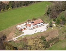 Veduta aerea di una casa rurale isolata con tetto in tegole rosse e cortile pavimentato, affiancata da un annesso con autorimessa. L’edificio è circondato da prati verdi e bosco su un pendio collinare, con strada sterrata di accesso.