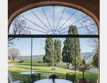 Grande finestra ad arco con cornice in mattoni e inferriata decorativa, che si affaccia su un giardino con prato curato. All’esterno si vedono alti cipressi, un muretto e colline sullo sfondo sotto un cielo azzurro.