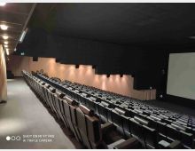 Interno di una sala cinematografica con lunghe file di poltrone grigie disposte in pendenza verso un grande schermo sul lato destro. Pareti scure con pannelli illuminati e corridoio laterale con luci a soffitto lungo il lato sinistro.