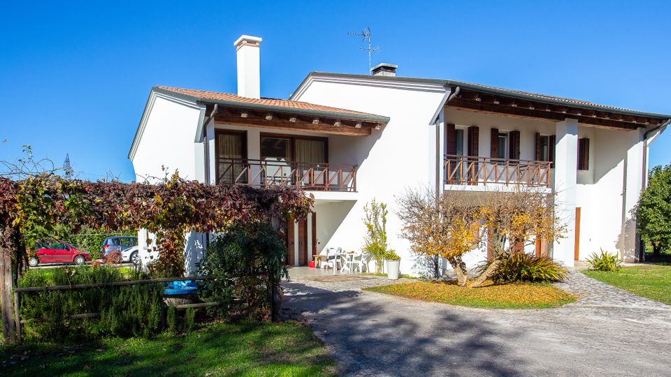 Casa indipendente a due piani con facciata bianca, tetto a falde e balconi in legno, affacciata su un cortile con vialetto in ghiaia. Davanti si vedono giardino con prato, una pergola coperta di rampicanti e alberi ornamentali sotto un cielo sereno.