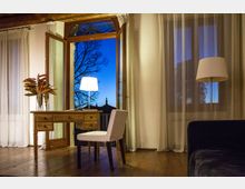 Interno di una stanza con pavimento in legno, scrivania in legno e sedia imbottita davanti a una grande porta-finestra ad arco aperta su un balcone, con cielo serale visibile all’esterno. Tende leggere alle finestre e lampade a stelo e da tavolo illuminano l’ambiente, con un divano scuro sul lato destro.