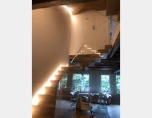 Interno moderno con scala sospesa in legno e vetro illuminata da strisce LED lungo i gradini, sotto un soffitto alto con travi a vista. Al piano inferiore si vede un’area soggiorno con poltrone e grandi finestre che affacciano sul verde esterno.