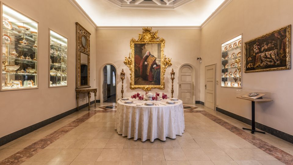 Sala museale in stile classico con pavimento in pietra e soffitto a cassettoni illuminato, con un grande ritratto in cornice dorata sulla parete di fondo. Al centro c’è un tavolo rotondo apparecchiato, mentre lungo le pareti laterali si vedono teche illuminate con porcellane e un grande specchio decorato.