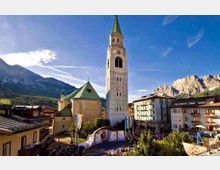 Piazza di un paese alpino con una chiesa in pietra e alto campanile con guglia verde, affiancata da edifici residenziali con balconi. Sullo sfondo si vedono montagne rocciose illuminate dal sole sotto un cielo azzurro.