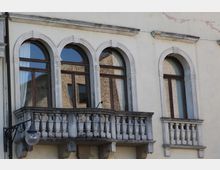 Facciata di un edificio storico con quattro alte finestre ad arco incorniciate in pietra e infissi in legno scuro. Al centro sporge un balcone in pietra con balaustra a colonnine, affiancato da un secondo balconcino più piccolo sulla destra.