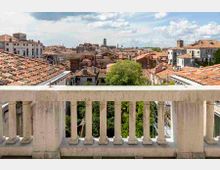 Vista panoramica da una terrazza con balaustra in pietra su un centro storico con numerosi tetti in coppi rossi e palazzi compatti. Sullo sfondo si distinguono campanili e torri sotto un cielo parzialmente nuvoloso, con alberi verdi tra gli edifici.