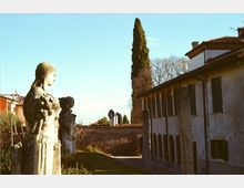 Cortile storico con statue in pietra in primo piano e un edificio in muratura con portico ad archi e tetto in coppi sul lato destro. Sullo sfondo si vedono un alto cipresso, un muro in mattoni e un vialetto lastricato sotto un cielo sereno.