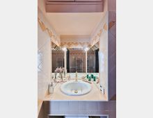 Angolo lavabo di un bagno con ampio specchio incassato, lavandino rotondo e rubinetteria dorata su piano piastrellato chiaro. Pareti rivestite in piastrelle con motivo geometrico e luci laterali, con accessori da toilette e asciugamano sotto il piano.