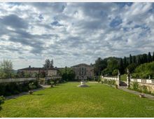 Ampio giardino formale con prato centrale e un grande vaso ornamentale su piedistallo, affiancato da viali in pietra, muretti e balaustre con statue. Sullo sfondo si vede una villa storica con facciata simmetrica e colline alberate sotto un cielo nuvoloso.