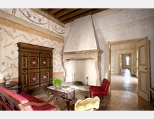 Salone storico con grande camino in pietra scolpita e cappa alta, soffitto con travi in legno e pareti decorate a motivi affrescati. Arredi d’epoca con divani in velluto rosso, tavolino centrale e un grande armadio intarsiato; una porta aperta conduce a un corridoio luminoso con pavimento in terrazzo.
