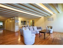 Soggiorno open space con pavimento in parquet e soffitto a travi in legno a vista dipinte di chiaro, con pareti color crema. Al centro ci sono un divano e poltrone chiare attorno a un tavolino, con zona pranzo e corridoio sullo sfondo.
