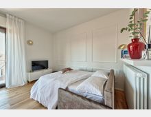 Camera da letto luminosa con pareti bianche a riquadri decorativi, letto matrimoniale con coperta bianca e pavimento in legno. A sinistra una grande finestra con tende leggere e un mobile basso con TV, a destra una mensola sopra il termosifone con libri e un vaso rosso con pianta.