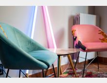 Angolo lounge interno con due poltrone imbottite (una verde e una rosa) disposte attorno a un tavolino basso con piano in vetro e gambe metalliche. Sullo sfondo si vedono tubi luminosi al neon appoggiati alla parete, un termosifone e un tappeto multicolore su pavimento in piastrelle.